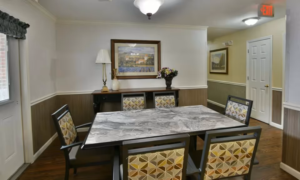 A dining room with a rectangular marble-patterned table surrounded by six chairs with geometric patterned upholstery. There is a side table against the wall with a lamp, a vase, and a flower arrangement. Two framed paintings hang on the walls, and the room has wood paneling on the lower half of the walls and light-colored paint above. A hallway with a white door and an exit sign is visible in the background.