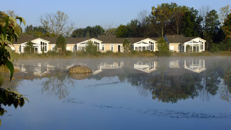 Row of single-story residential buildings with large windows, reflected in a calm body of water with some mist rising above the surface, surrounded by trees and greenery under a clear sky.