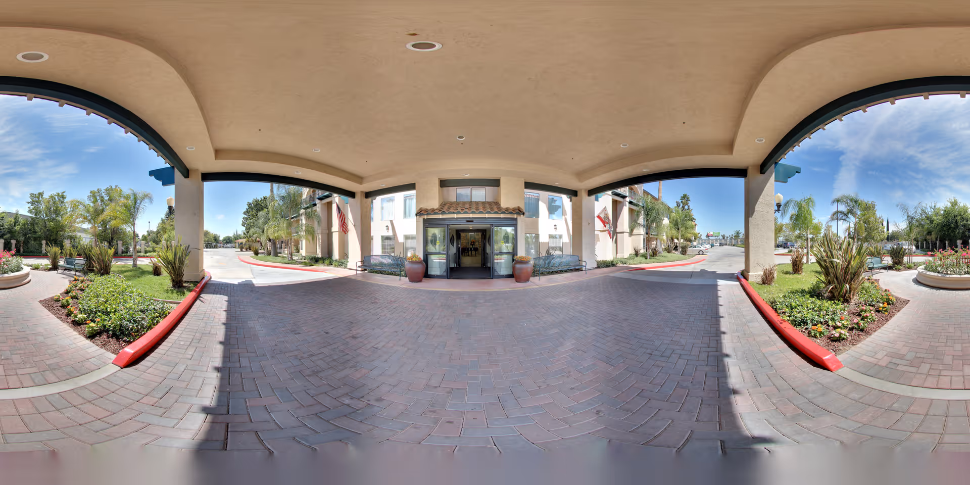 Covered entrance area of a building with a paved driveway and landscaped garden beds on either side. The building has large glass doors at the entrance, two large potted plants, and American flags displayed. The sky is clear with some clouds, and palm trees and other greenery are visible around the entrance.