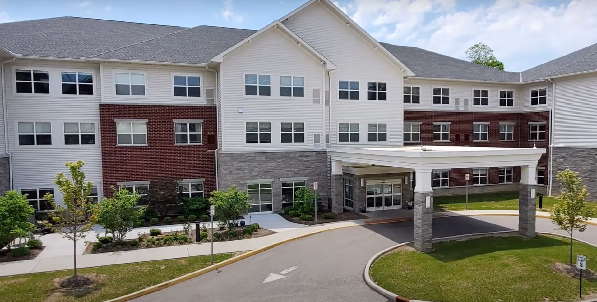 Exterior view of a three-story senior living facility building with a covered entrance, multiple windows, and landscaped greenery including small trees and shrubs. The driveway curves in front of the entrance with a speed limit sign visible on the grass.