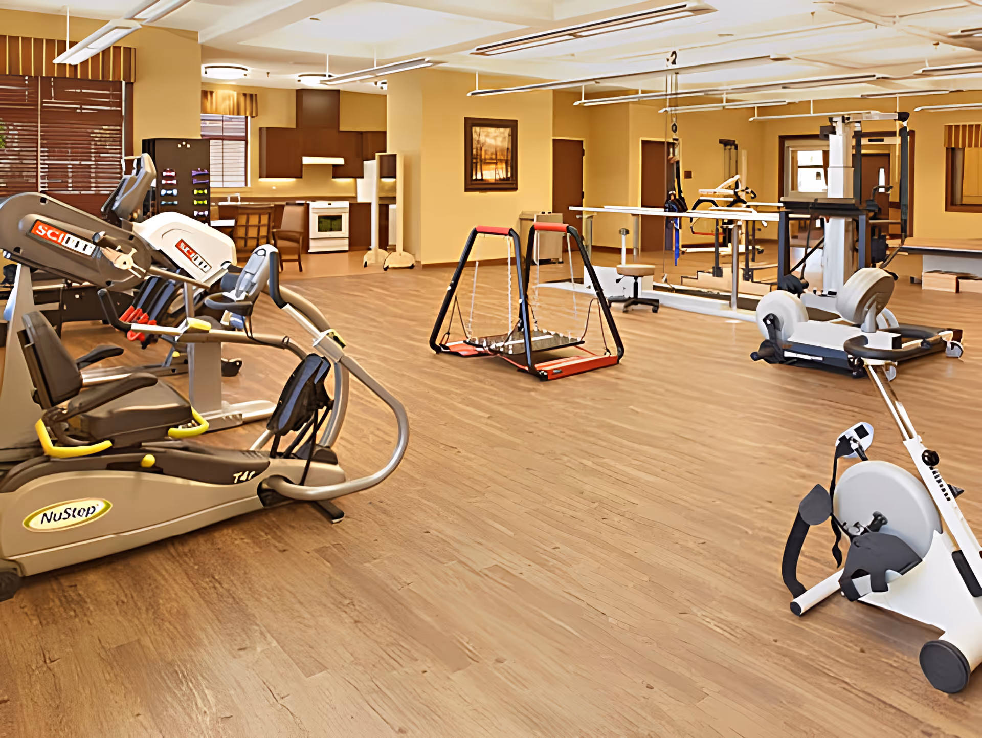 A spacious rehabilitation or physical therapy room with exercise equipment including stationary bikes, a swing, parallel bars, and other therapy apparatus. The room has wooden flooring, beige walls, and a small kitchen area in the background with a stove and cabinets. Large windows with blinds allow natural light to enter.
