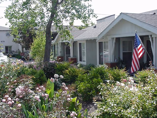 Exterior view of a single-story residential building with a garden full of blooming flowers and shrubs in front. An American flag is displayed near the entrance of the building. Trees and other buildings are visible in the background under a clear sky.