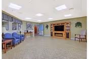 Bright common lounge with chairs, small tables, and a large wooden TV/entertainment center.
