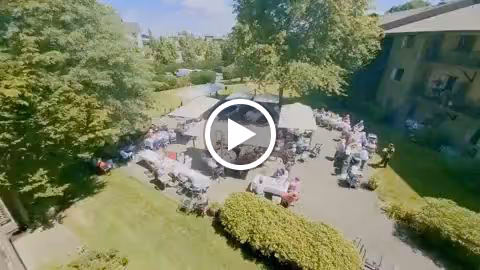 Aerial view of an outdoor gathering at Willamette Oaks with people seated at tables under umbrellas and a tent on a paved patio area surrounded by trees and greenery, adjacent to a multi-story building.