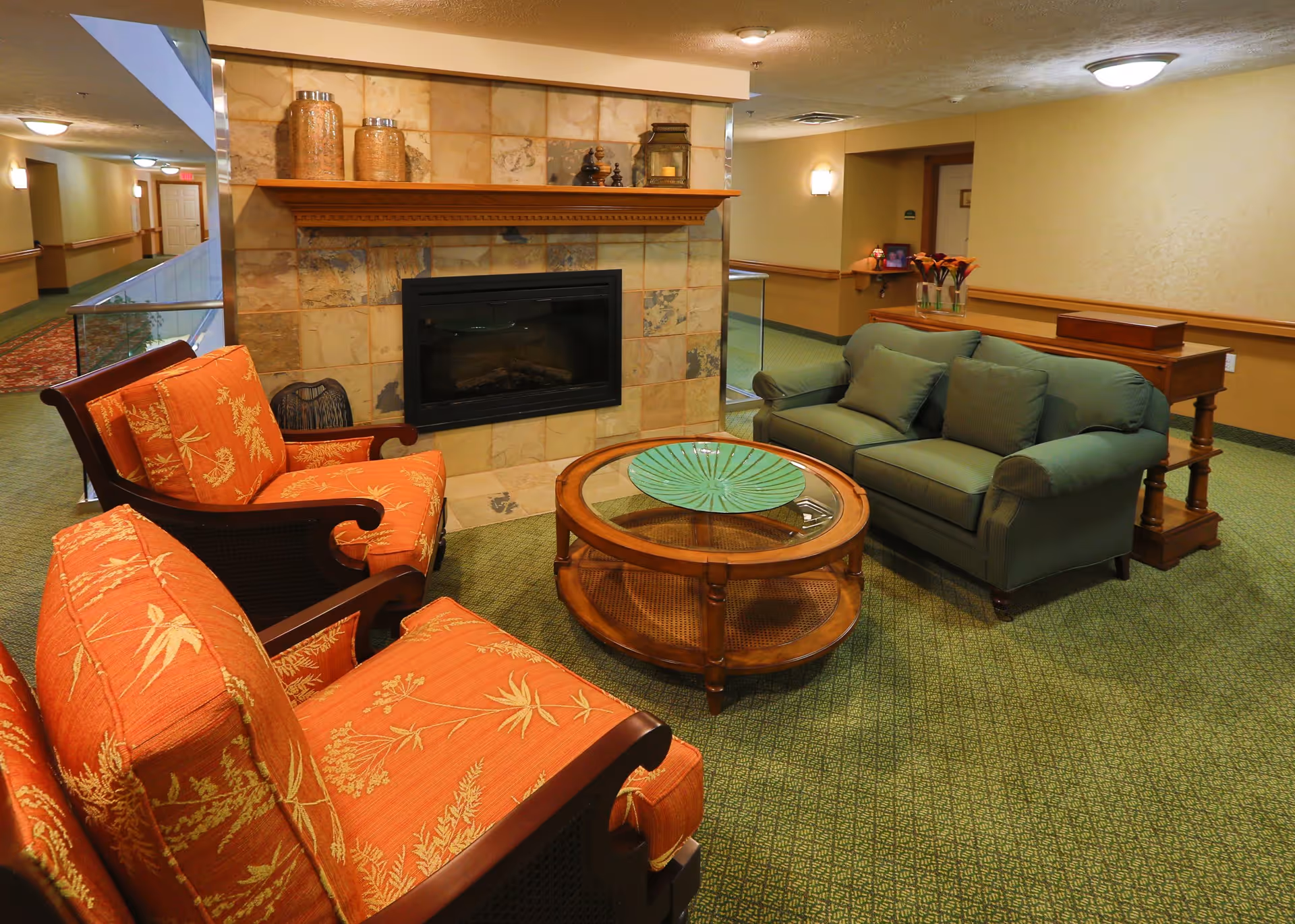 A cozy seating area in a retirement facility featuring two orange cushioned armchairs with floral patterns, a green cushioned sofa with three pillows, a round wooden coffee table with a decorative green plate, and a fireplace with a stone surround and wooden mantel. The room has green carpet and beige walls with soft lighting.
