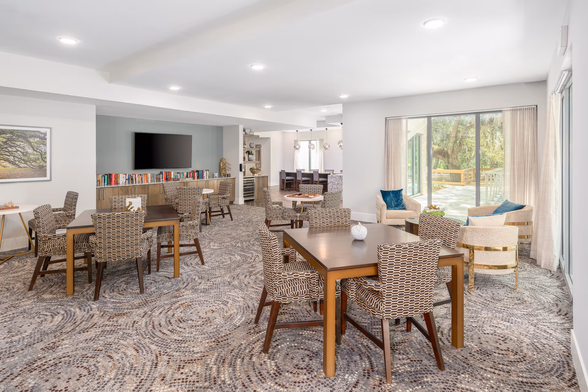 A bright and spacious common area in a senior living facility featuring multiple tables with patterned chairs, a seating area with armchairs and blue cushions near large sliding glass doors that open to an outdoor patio. A wall-mounted TV is above a cabinet filled with books, and the room has modern lighting and a patterned carpet.