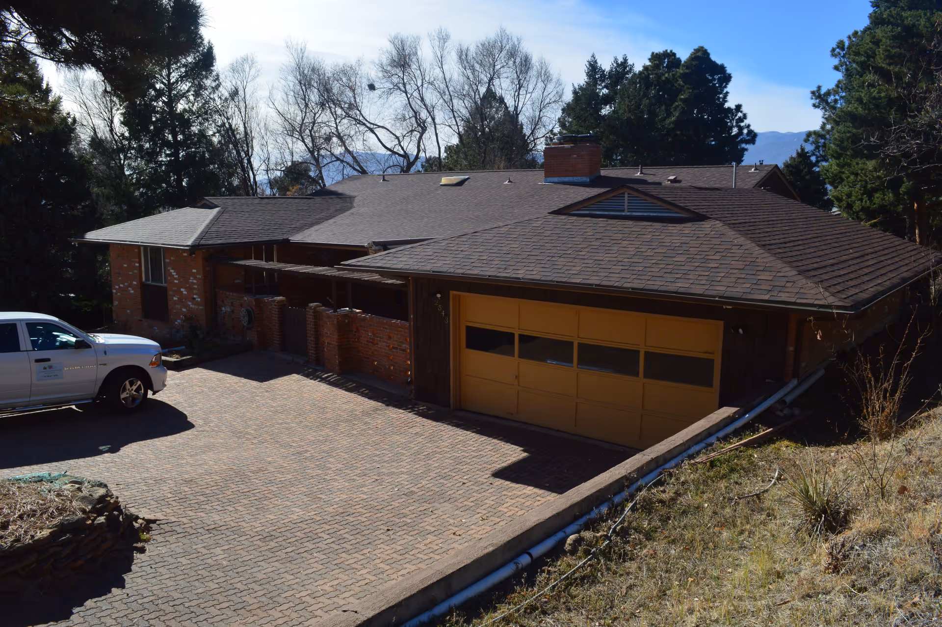 Exterior view of a single-story brick building with a brown shingled roof and a yellow garage door. A white pickup truck is parked on the paved driveway in front of the building. Trees and mountains are visible in the background under a clear blue sky.