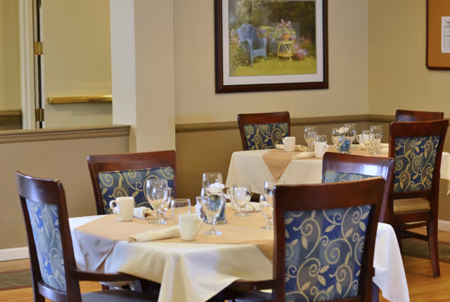 Small dining room with round tables set with glasses, cups, and napkins and patterned wooden chairs.