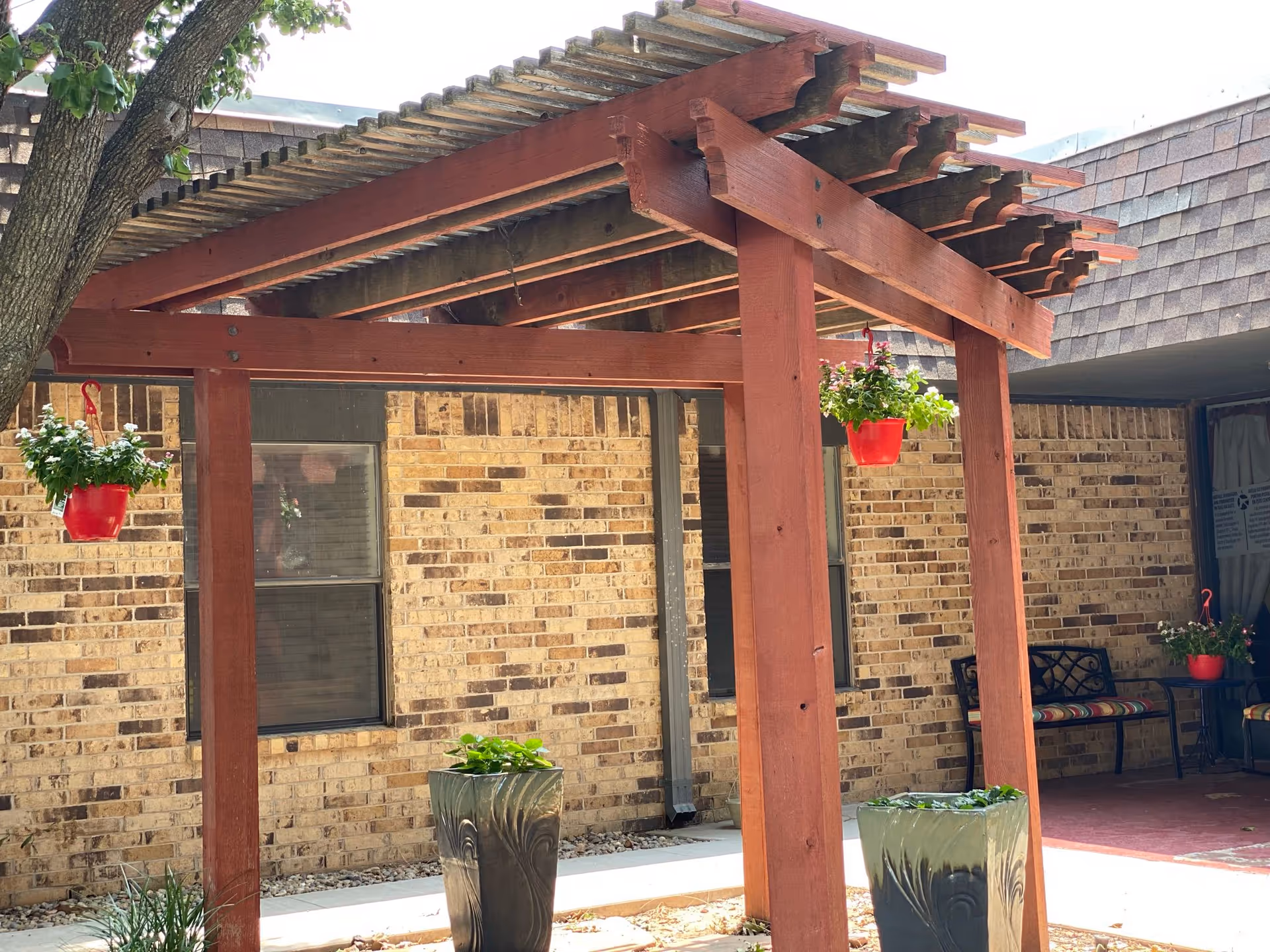 Outdoor seating area at Midwestern Healthcare Center featuring a wooden pergola with hanging red flower pots, two large planters with greenery, a brick wall background, and a bench with colorful cushions under a covered patio.