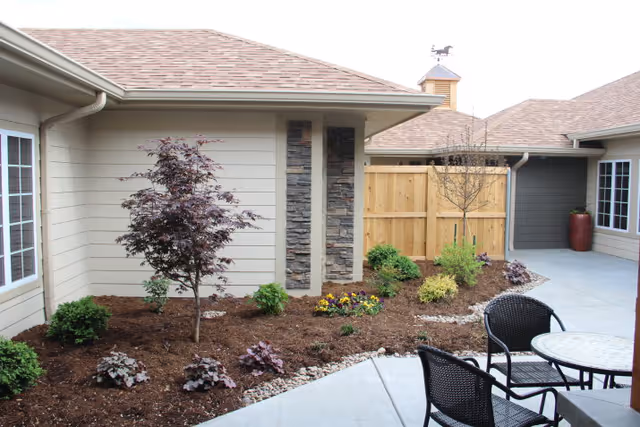 Outdoor patio area at ComfortCare Homes Kansas featuring a small garden with various plants and shrubs, a wooden privacy fence, and a round table with black wicker chairs on a concrete surface.