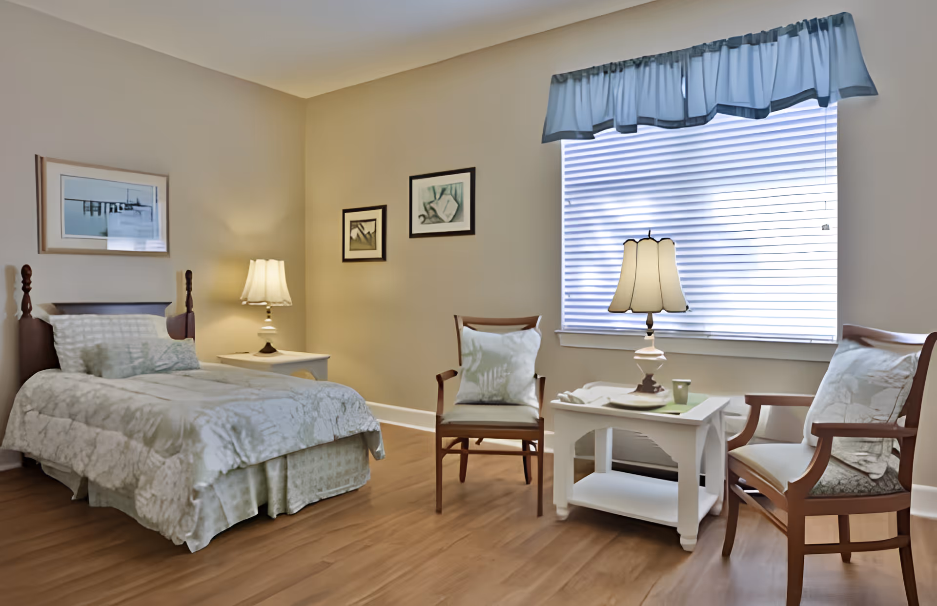 A cozy bedroom in a senior living facility with a single bed covered in light-colored bedding, a nightstand with a lamp, two wooden chairs with cushions, a small white table with a lamp and a cup, and three framed pictures on the beige walls. A window with blinds and a blue valance lets in natural light.