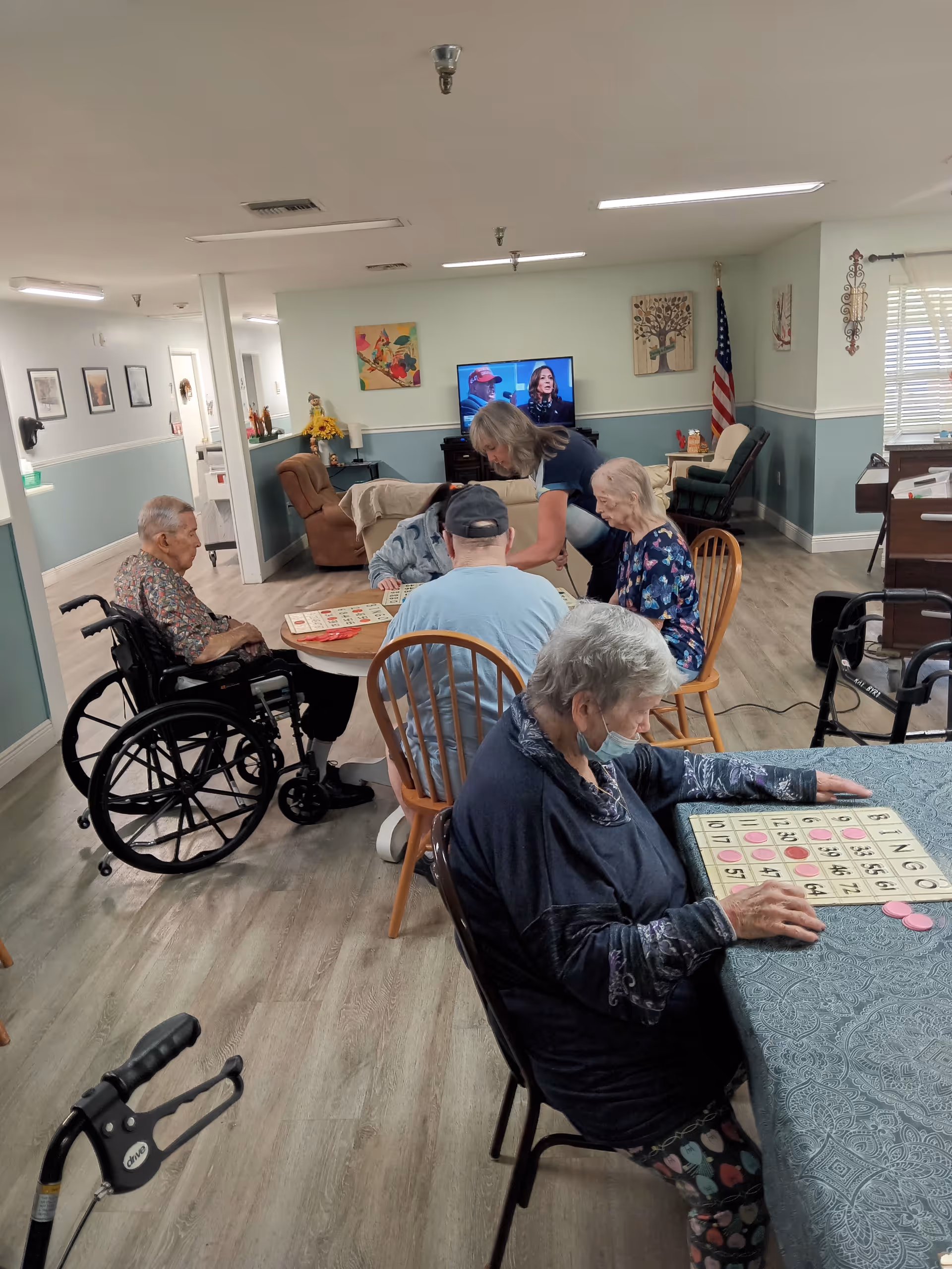 A group of elderly people sitting around tables in a common room playing bingo. One woman is placing bingo chips on her card, while others are engaged in the game. A caregiver is assisting one of the players. The room has light green walls, wooden flooring, and a television in the background. An American flag and various decorations are visible on the walls.