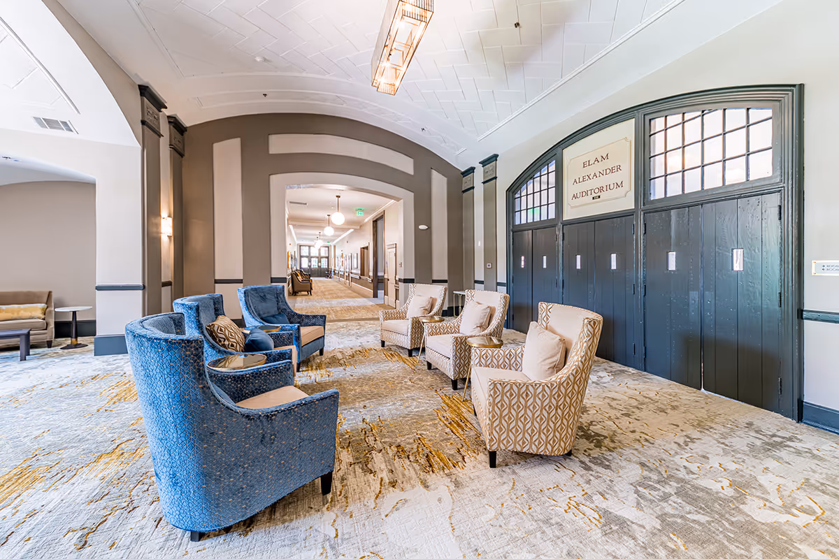 A spacious and well-lit lounge area with a mix of blue and beige upholstered armchairs arranged in small groups on a patterned carpet. The room features arched doorways and a sign above dark wooden doors reading 'Elam Alexander Auditorium'. The walls are painted in neutral tones with decorative molding, and the ceiling has a geometric pattern with a modern light fixture.