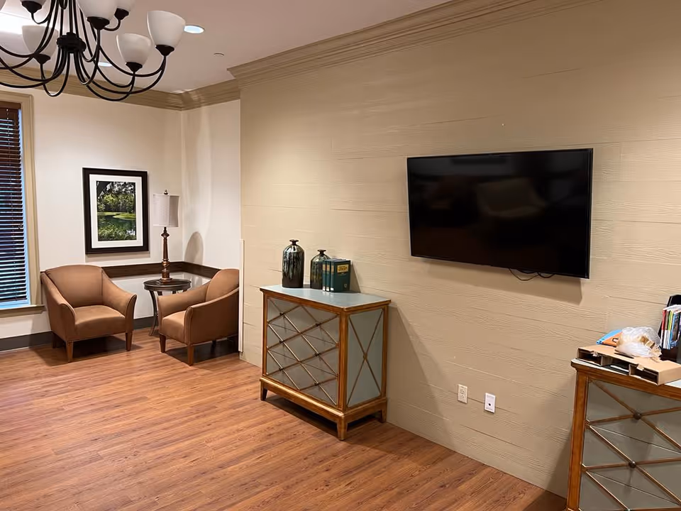 A lounge area with two upholstered chairs and a small table by a window, a wall-mounted TV, decorative cabinets, and hardwood floors.