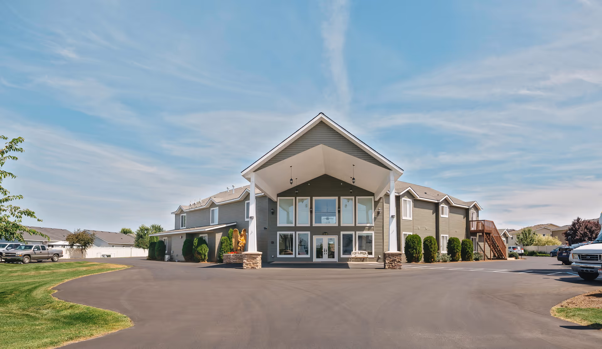 Front exterior view of a two-story senior living facility building with a large covered entrance, multiple windows, and surrounding parking area under a partly cloudy sky.