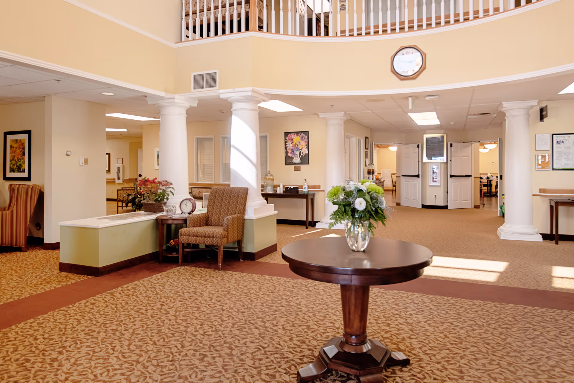 A spacious and well-lit common area in a senior living facility with beige walls and carpeted floors. The room features white columns, a round wooden table with a vase of flowers in the center, and comfortable upholstered chairs. There are framed pictures on the walls, a clock above the entrance, and double doors leading to another room.