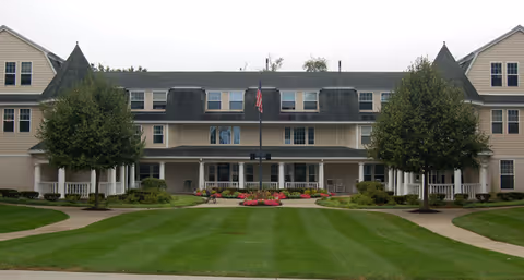 Front exterior view of a large, beige senior living facility building with multiple windows, two symmetrical trees, a well-maintained lawn, and an American flag in the center.