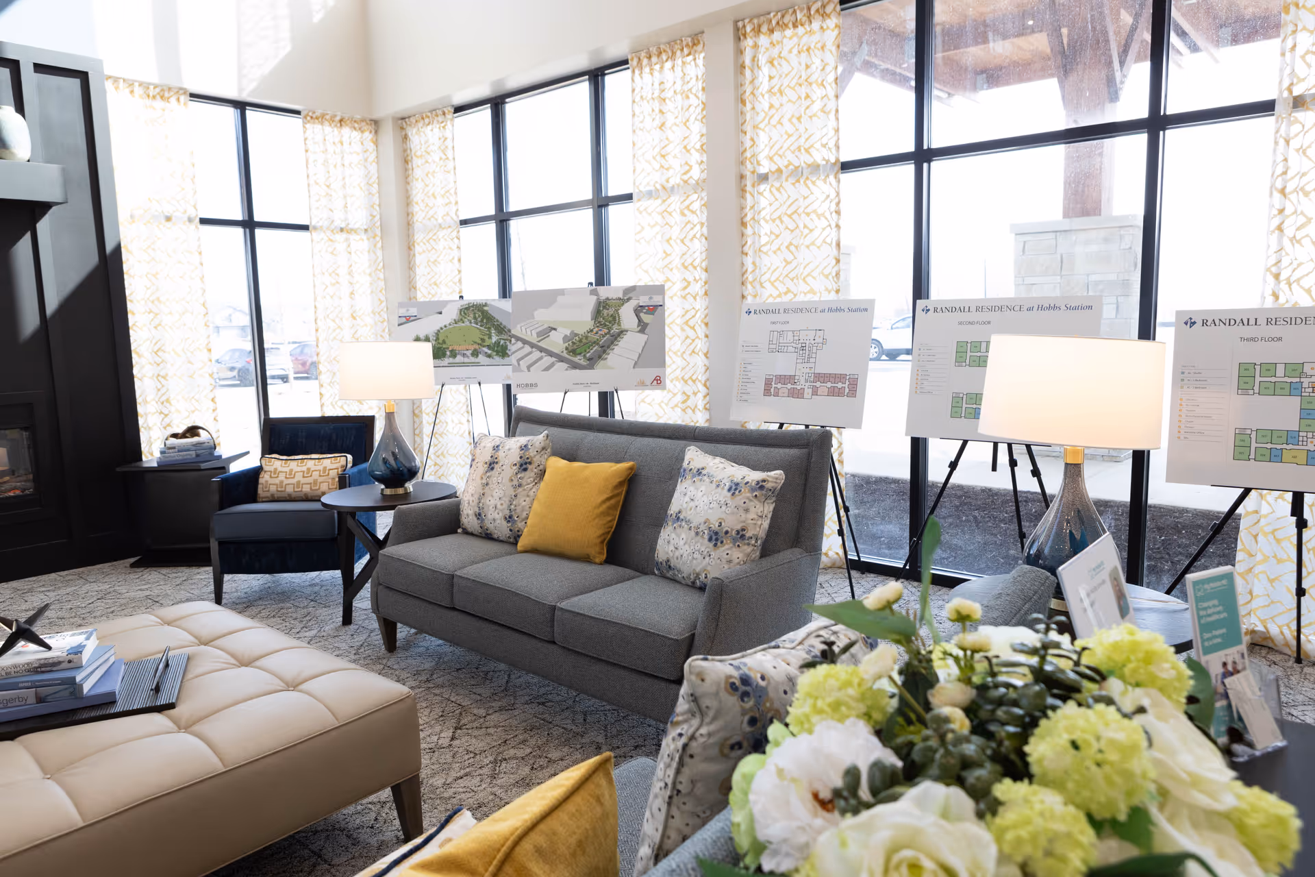 A bright and modern living room area with large windows covered by yellow patterned curtains. The room features a gray sofa with decorative pillows, a blue armchair, a beige tufted ottoman, and two table lamps with blue bases. Behind the sofa, there are several easels displaying floor plans and site maps for Randall Residence at Hobbs Station. A floral arrangement is visible in the foreground.