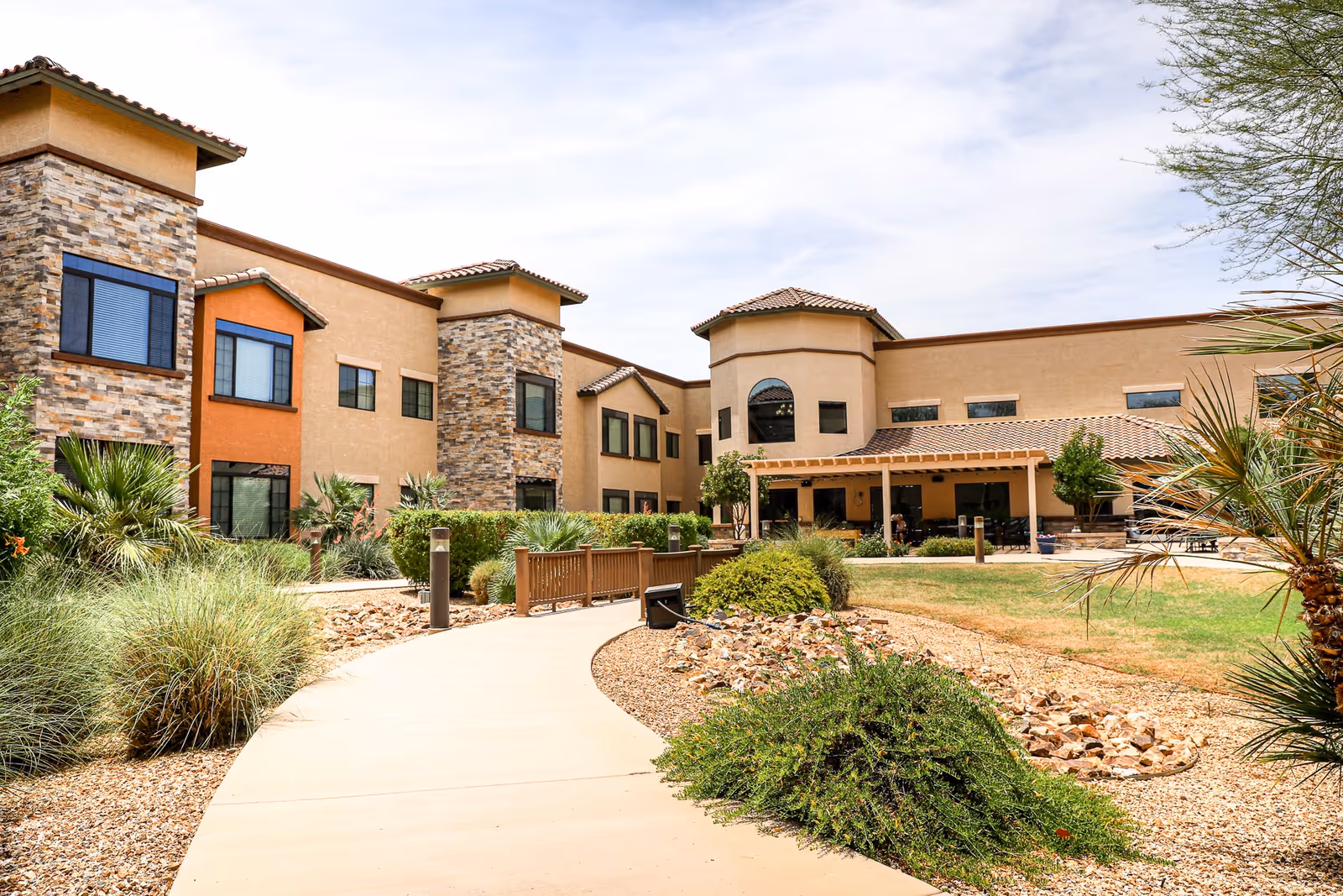 Exterior view of Orchard Pointe at Surprise, showing a two-story building with a mix of stone and stucco facade, surrounded by landscaped desert plants and a paved walkway leading to the entrance under a covered patio.