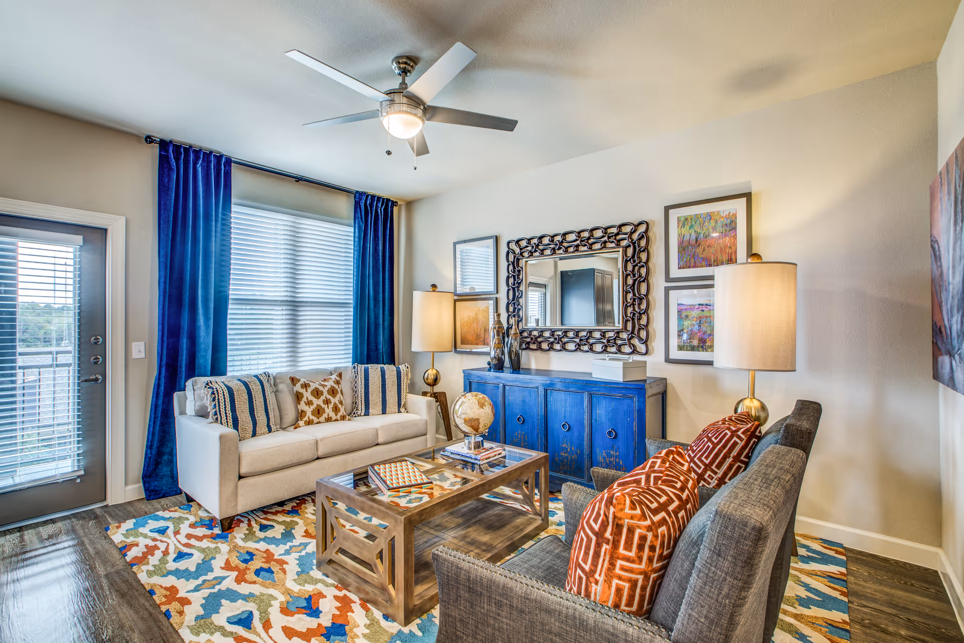 A bright living room with a beige sofa adorned with patterned pillows, a gray armchair with orange geometric pillows, a wooden coffee table with books and a globe, a blue sideboard with a decorative mirror above it, two table lamps with beige shades, colorful abstract paintings on the walls, blue curtains framing a large window, and a ceiling fan with a light.