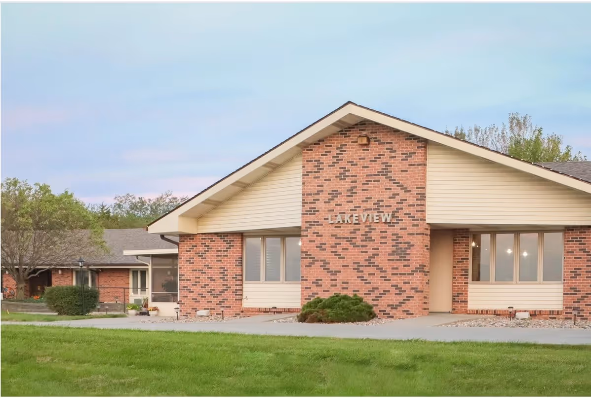 Exterior view of a single-story brick building with a peaked roof and the word 'LAKEVIEW' displayed on the front brick wall. The building is surrounded by green grass and some trees in the background under a clear sky.
