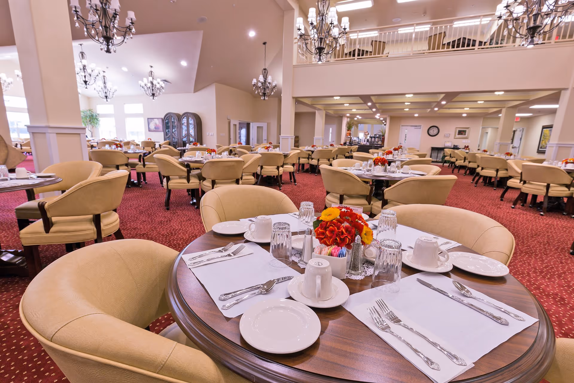 A spacious dining room with multiple round tables set with white plates, cups, silverware, and glasses. Each table has a centerpiece of red and yellow flowers. The room features beige upholstered chairs, red carpet with a subtle pattern, chandeliers hanging from the ceiling, and a second-floor balcony overlooking the area.