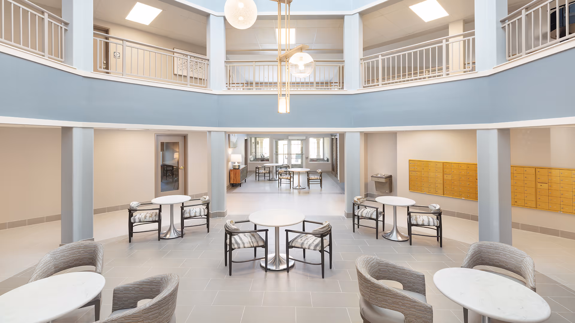 A spacious, modern common area with multiple round tables and chairs arranged for seating. The room features a two-story design with a balcony railing on the upper level. There are yellow mailboxes mounted on the wall to the right and a water fountain beneath them. The area is well-lit with natural light coming through glass doors and windows at the far end, and pendant lights hanging from the ceiling.