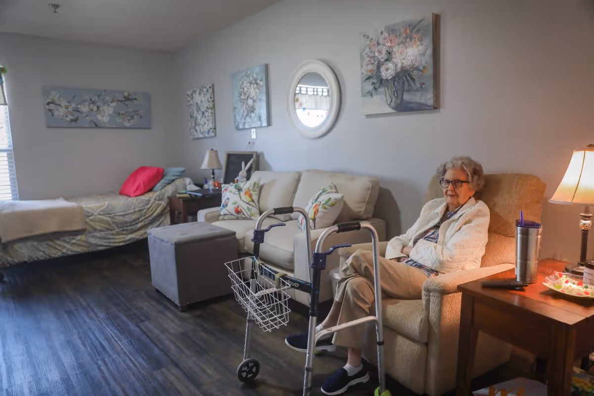 An elderly woman sitting in a beige armchair in a cozy room with a bed, a sofa, and a walker in front of her. The room has wooden flooring, floral paintings on the wall, a small side table with a lamp and a tumbler, and a window with blinds letting in natural light.