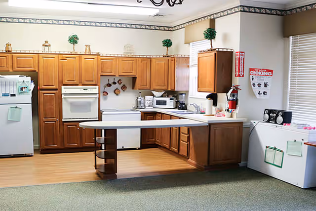 A kitchen area in a senior living facility featuring wooden cabinets, a white refrigerator, built-in oven, microwave, dishwasher, and a countertop with an extended table. The kitchen has various small appliances, a fire extinguisher, and a choking emergency poster on the wall. There are also decorative plants on top of the cabinets and a window with blinds.