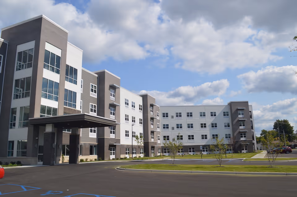 Modern four-story senior living building with a covered entrance, parking lot, and lawn under a partly cloudy sky.
