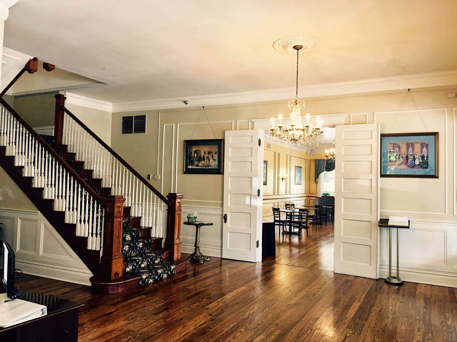 Interior view of a senior living facility showing a wooden staircase with floral carpet runner on the steps, leading to an upper floor. The room has polished hardwood floors, beige walls with white trim, and two framed paintings on the walls. Double white doors open into a dining area with tables and chairs, illuminated by a chandelier hanging from the ceiling.