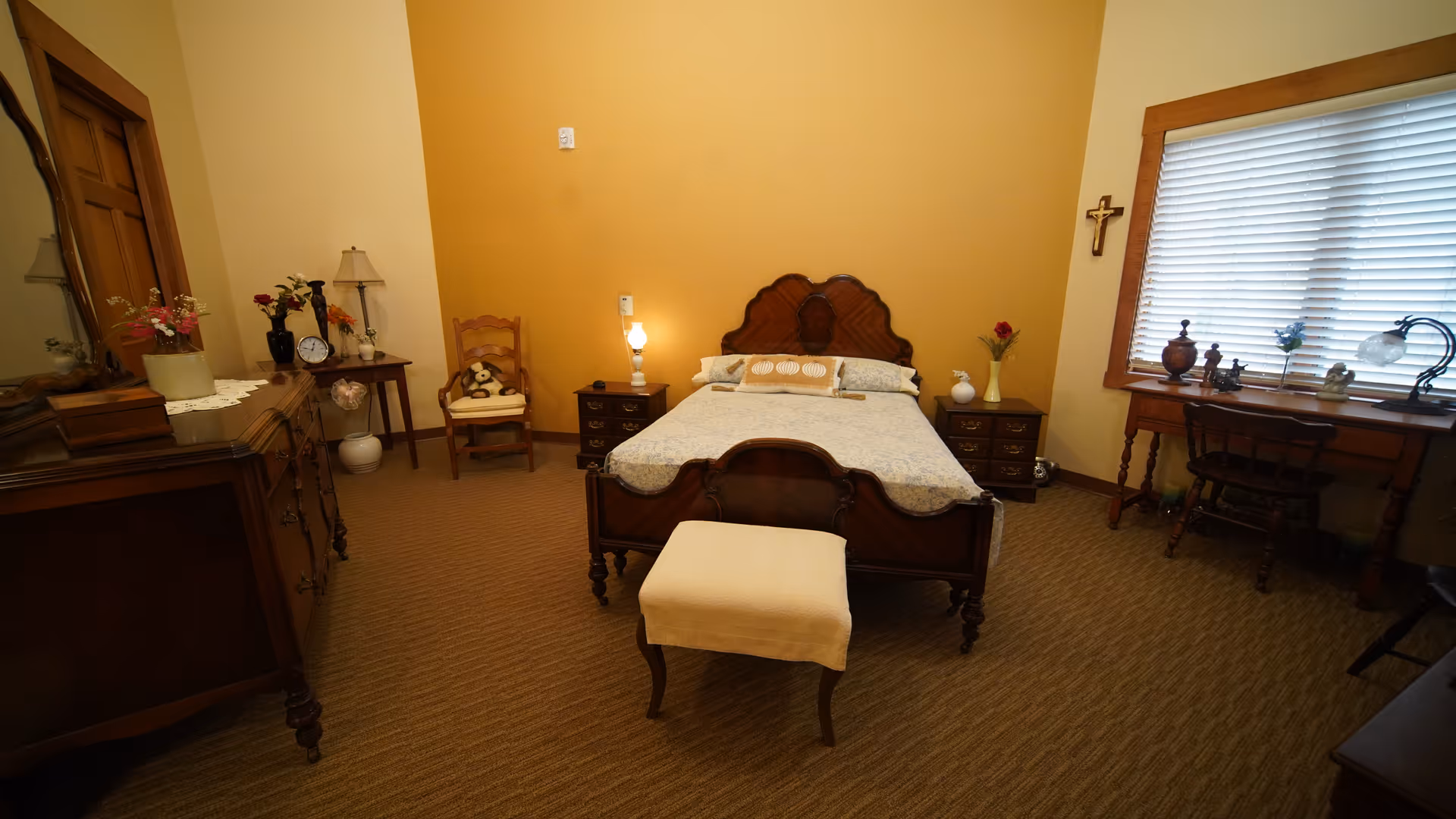 A cozy bedroom with a vintage wooden bed centered against a two-tone yellow and beige wall. The bed has a patterned bedspread and a small cushioned bench at its foot. On either side of the bed are matching wooden nightstands with lamps and decorative items. To the left, there is a wooden dresser with a mirror and flowers on top. A wooden chair with a stuffed animal sits in the corner. On the right side, a wooden desk with a chair is placed under a window with blinds, and a small cross hangs on the wall beside it.