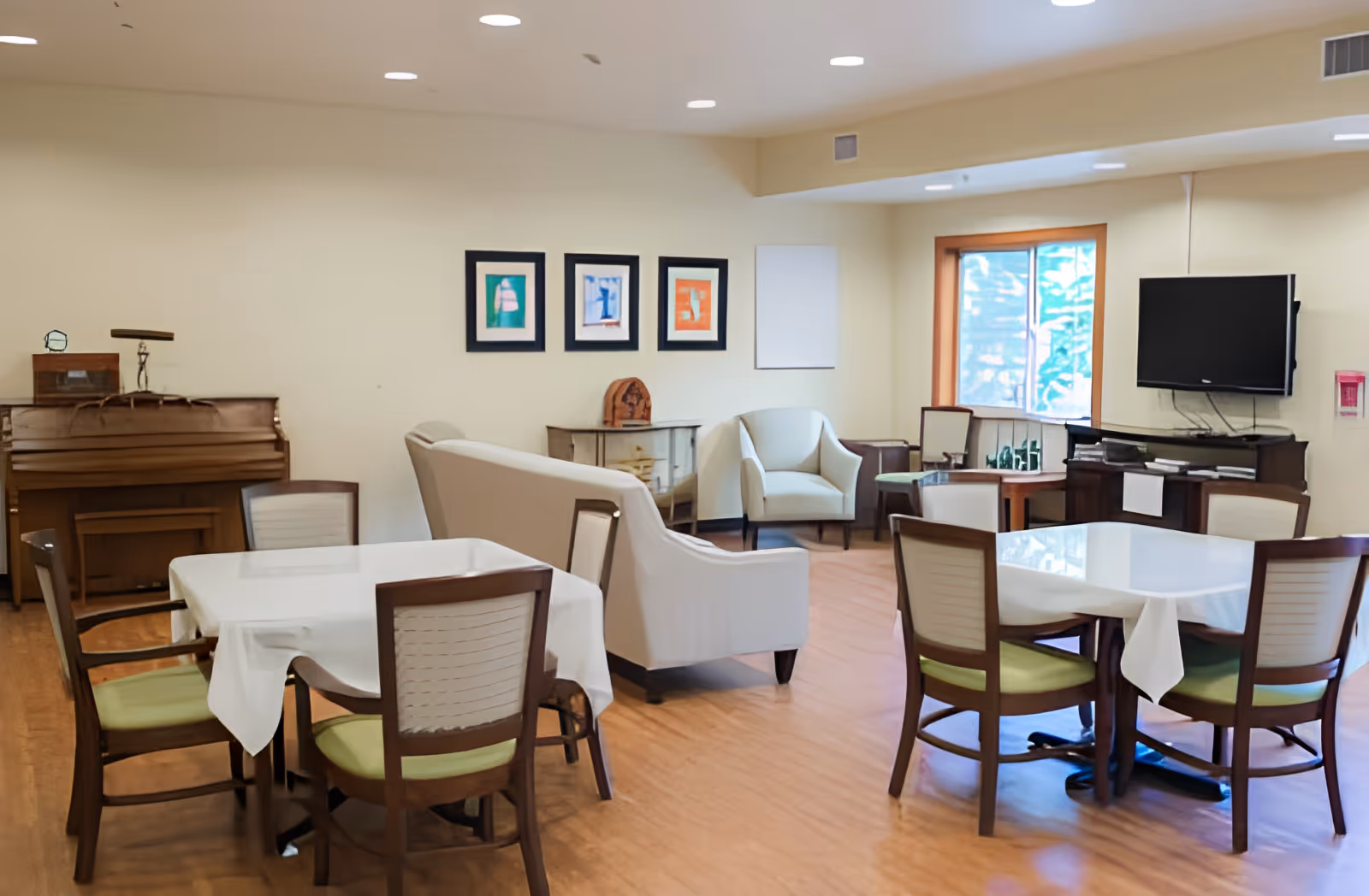 A cozy common area with two tables covered with white tablecloths, each surrounded by four wooden chairs with green cushions. There is a beige sofa and two armchairs arranged near a window and a wall-mounted TV. A piano is placed against the wall, and three colorful framed artworks hang above a small cabinet.