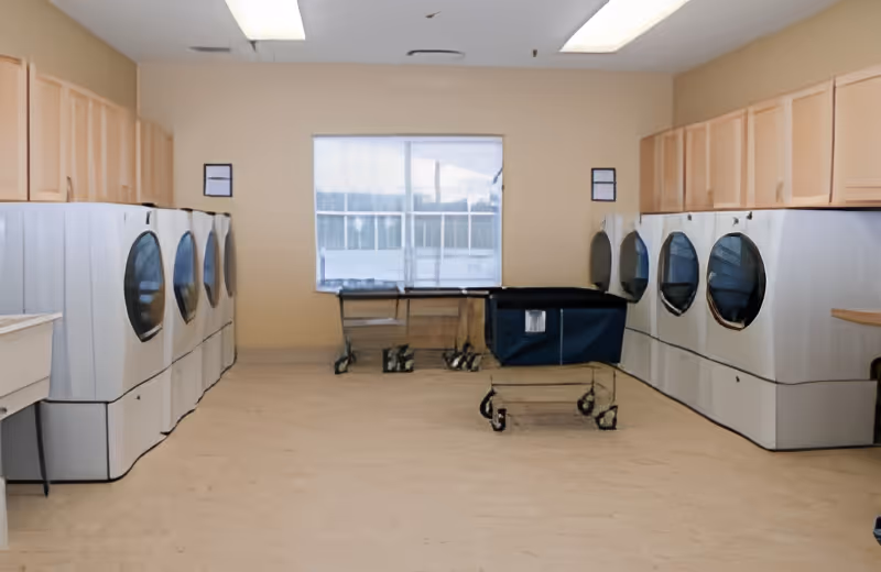 Laundry room with multiple front-loading washing machines and dryers lined up against two opposite walls, beige walls and cabinets above the machines, a window at the far end, and laundry carts in the center of the room.