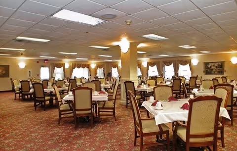 A spacious dining room with multiple tables covered in white tablecloths and set with cups and napkins. The room has patterned carpet flooring, upholstered chairs, large windows with curtains, and ceiling lights providing bright illumination.