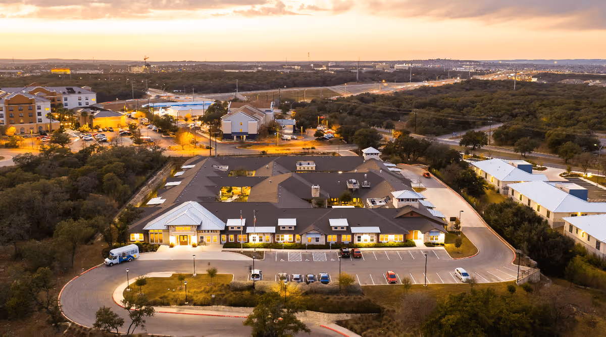 Aerial view of Waterford at Westover Hills senior living facility at dusk, showing the building with illuminated windows, surrounding parking lot with cars, nearby buildings, and a landscape of trees and hills in the background.