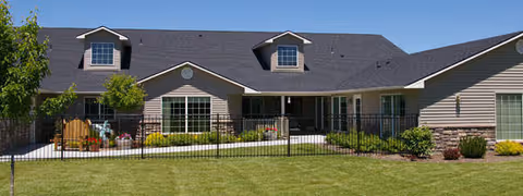 Single-story cottage-style assisted living building with dormer windows, a fenced patio and manicured lawn under a clear blue sky.