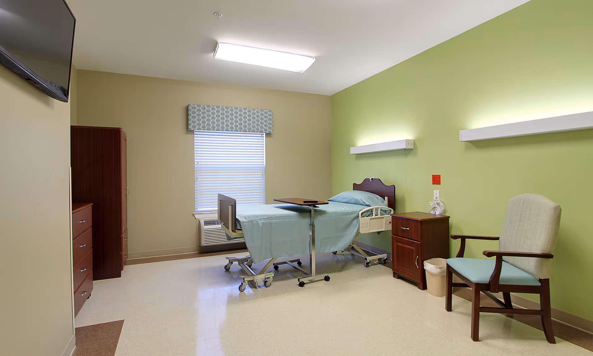 A clean and simple patient room with a hospital bed covered in light blue sheets, a wooden nightstand with a lamp, a comfortable armchair, a window with blinds and a patterned valance, and a wall-mounted TV. The walls are painted light green and beige.