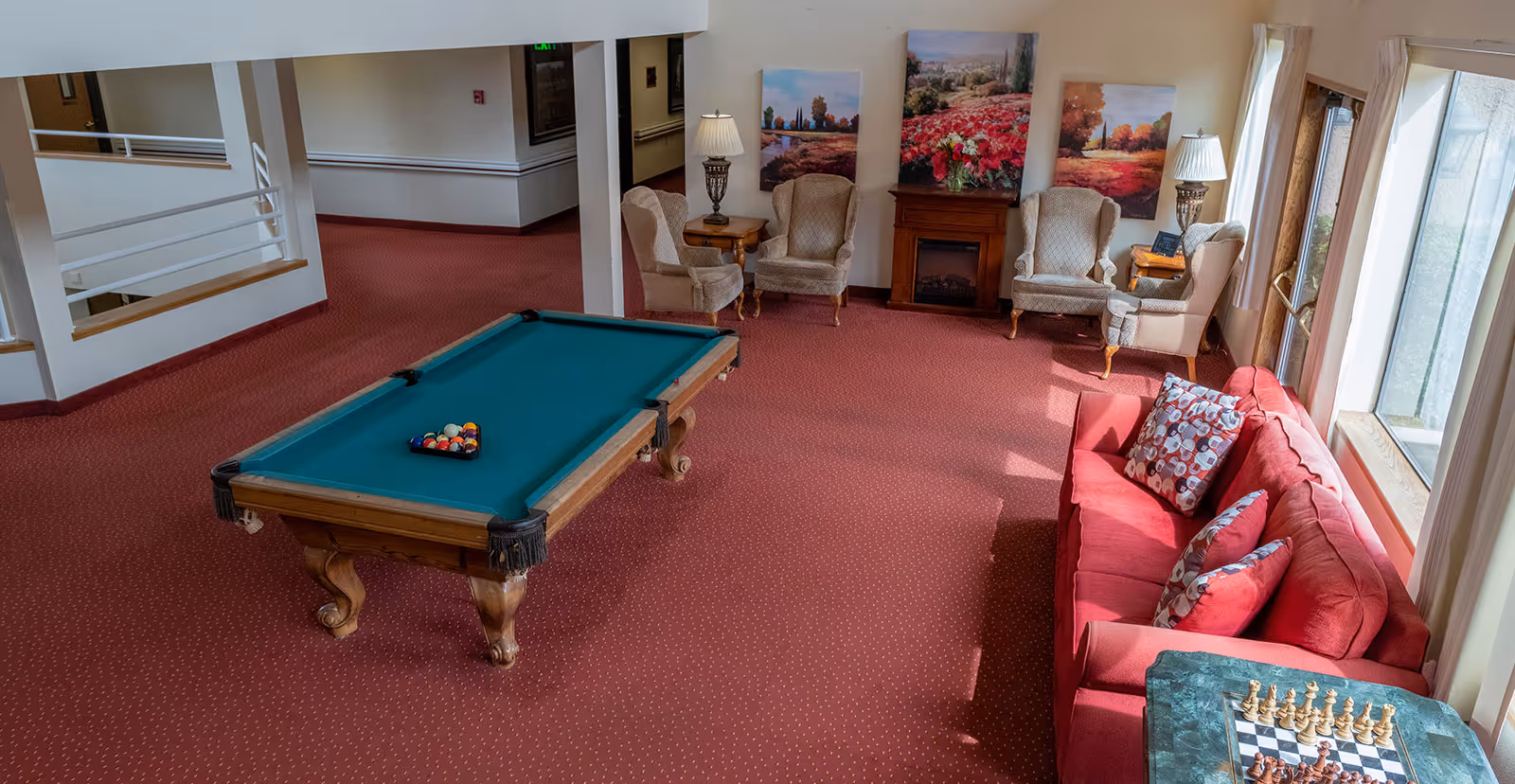 A cozy common area in a senior living facility featuring a pool table with balls arranged for a game, a red couch with patterned pillows, a chessboard on a small table, four upholstered armchairs arranged around a wooden electric fireplace, two table lamps, and three landscape paintings on the wall. The room has red carpet and large windows letting in natural light.
