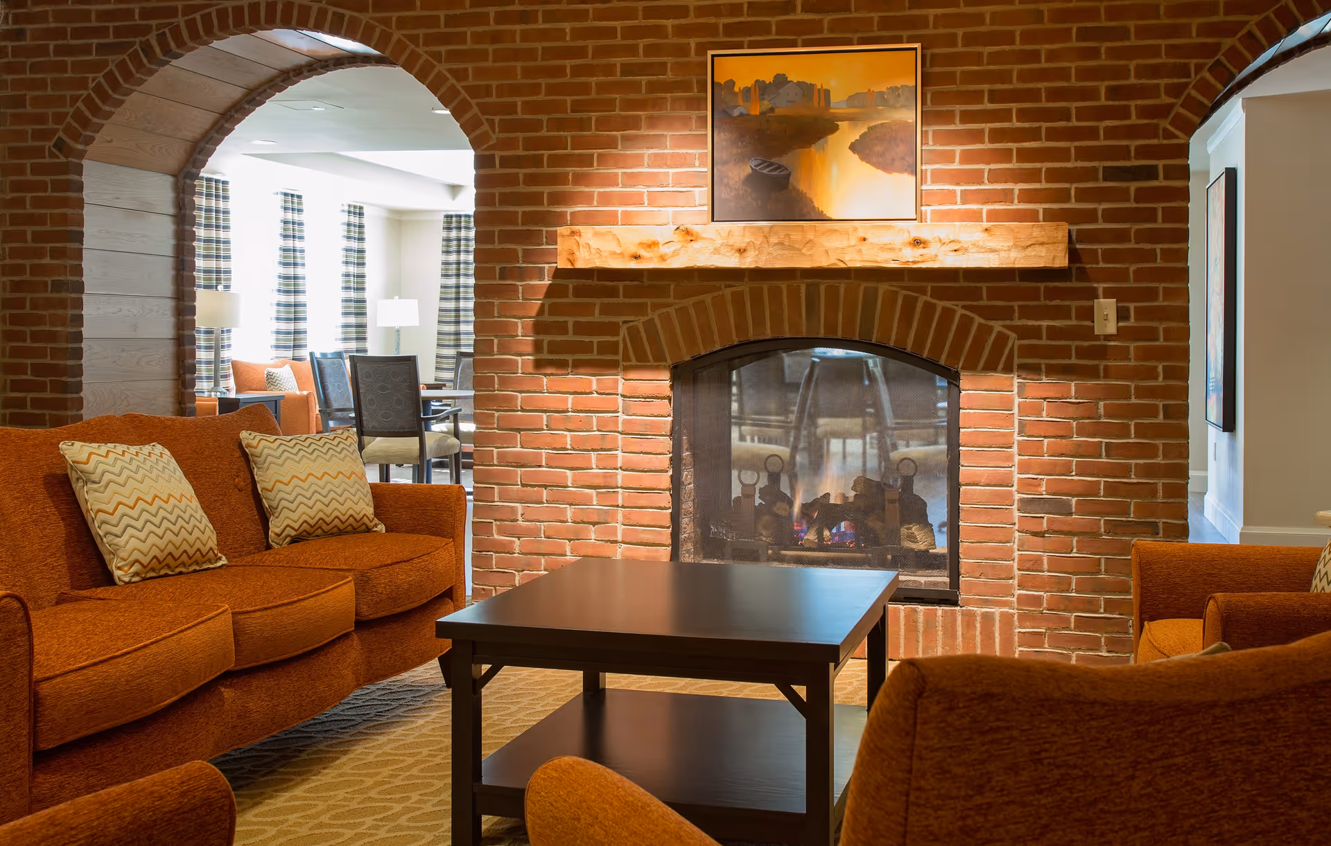 A cozy living room area with orange upholstered sofas and armchairs arranged around a dark wooden coffee table. The room features a brick fireplace with a wooden mantel and a painting of a river scene above it. In the background, there is a dining area with tables and chairs near windows with striped curtains.