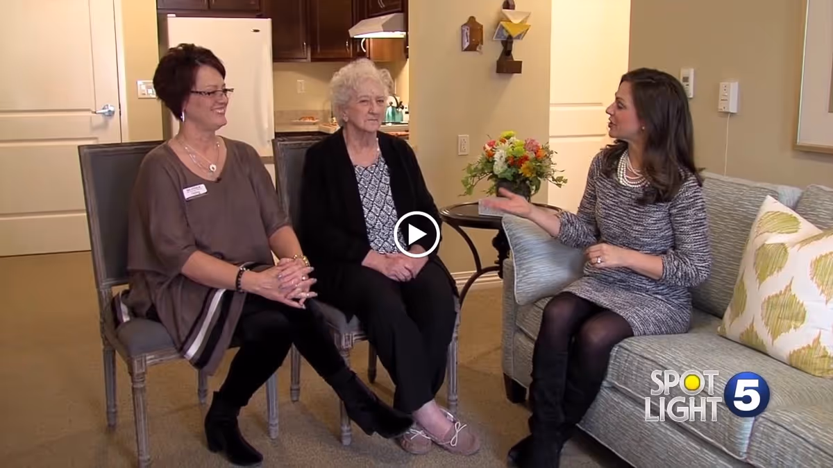 Three women sitting and talking in a cozy living room area with a kitchen visible in the background. Two women are seated on chairs, and one woman is sitting on a couch next to a small table with a flower arrangement.