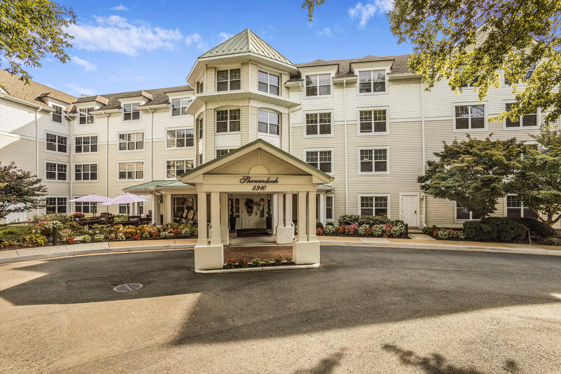 Exterior view of a multi-story senior living facility named Shenandoah with white siding and multiple windows. The entrance features a covered portico with the number 5910 and landscaped flower beds around the driveway. There are outdoor seating areas with umbrellas on the left side and trees framing the scene under a partly cloudy blue sky.