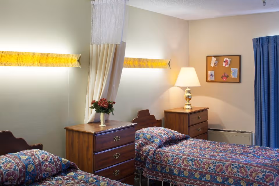 A bedroom with two single beds featuring floral patterned bedspreads. Between the beds is a wooden nightstand with a vase of flowers. Another wooden nightstand with a lamp is next to one of the beds. A curtain is partially drawn between the beds, and a corkboard with pinned notes is on the wall. Blue curtains cover a window on the right side.