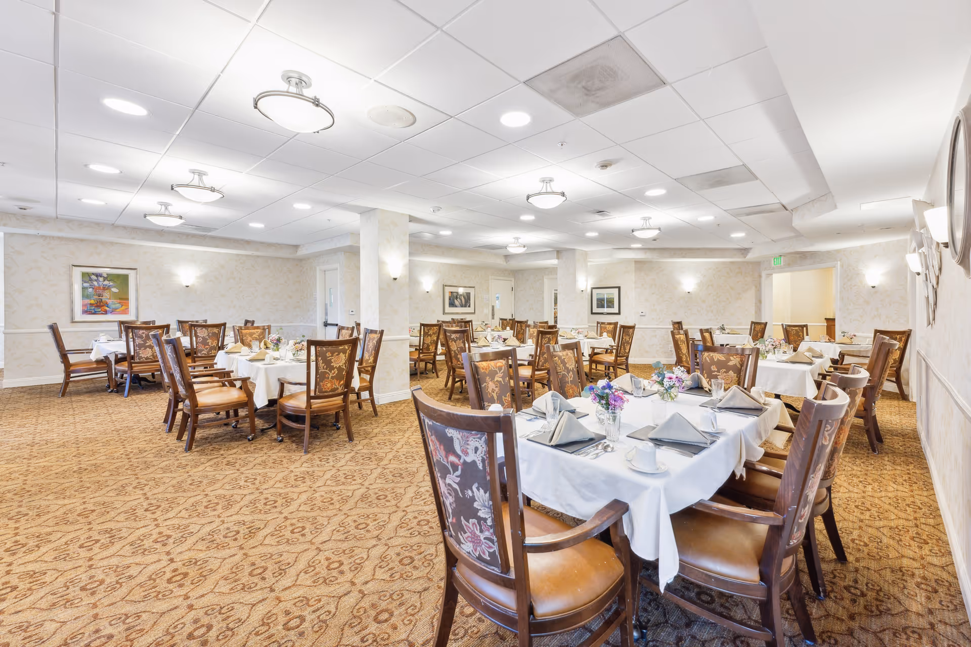 Spacious dining room with multiple tables set with white tablecloths, napkins, and floral centerpieces.