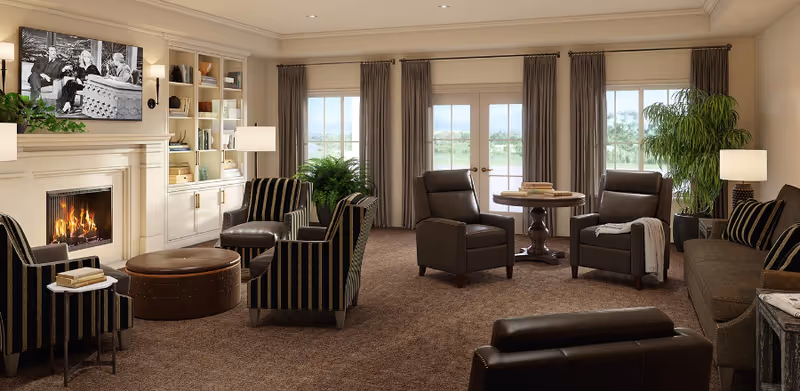 Comfortable senior living room with a fireplace, built-in bookshelves, multiple upholstered chairs and sofas, a round table, and French doors with drapes showing outdoor views.