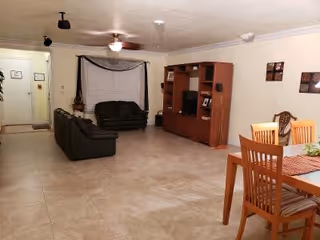 A spacious living room area with beige tiled flooring and cream-colored walls. The room features two dark green sofas arranged near a large wooden entertainment center with a TV and decorative items. To the right, there is a wooden dining table with four chairs and a table runner. The room is lit by ceiling lights and a ceiling fan, with a window covered by blinds and a valance.
