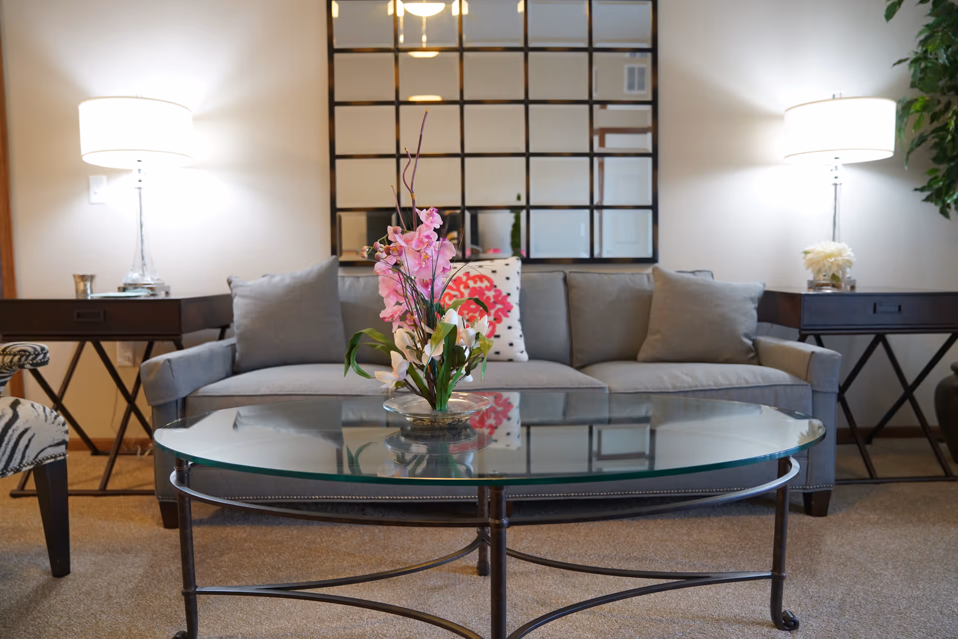 A cozy living room with a gray sofa adorned with cushions, a glass coffee table with a floral arrangement, two side tables each with a lamp, and a large decorative mirror on the wall behind the sofa.