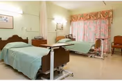 A hospital-style room with two beds, each covered with light blue sheets. The room has light-colored walls, a window with floral curtains, and medical equipment near the beds. There are two chairs near the window and a framed picture on the wall.
