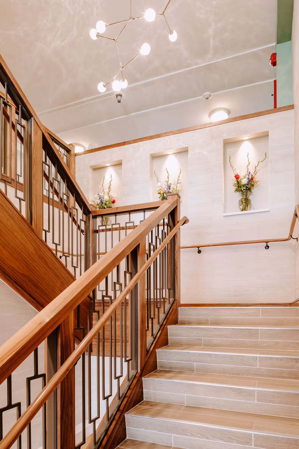 A well-lit indoor staircase with wooden handrails and metal balusters. The walls are light-colored with three recessed niches, each containing a vase with colorful flowers. A modern chandelier with multiple round bulbs hangs from the ceiling above the stairs.