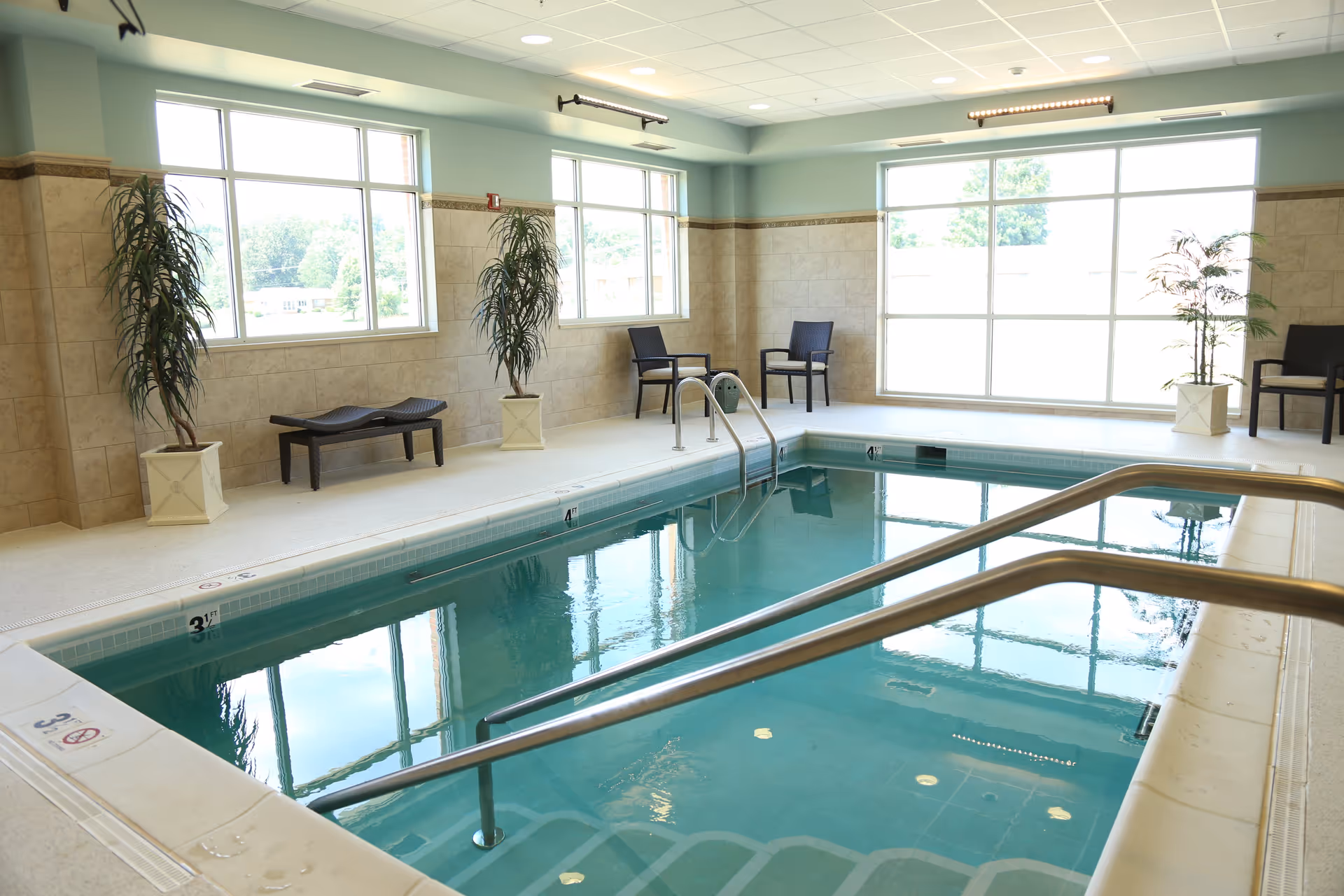 Bright indoor pool room with handrails, lounge chairs, potted plants and large windows in a senior living facility.